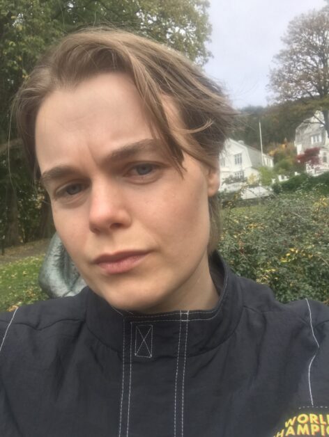 Selfie of white young person. Up close and underneight perspective. Wearing black jacket in green outdoor environment. Serious frowning face, short dark blond hair covering one eye. Photo by Oda Brekke.