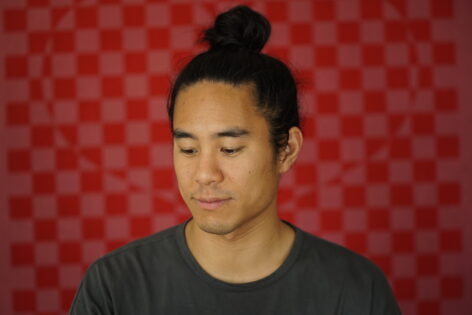 Tom's head is tilted down slightly, looking towards the lower left corner of the photo. Framed from his shoulders to his topknot, against an out-of-focus red-and-pink checkerboard background. Photo by Becca Green.