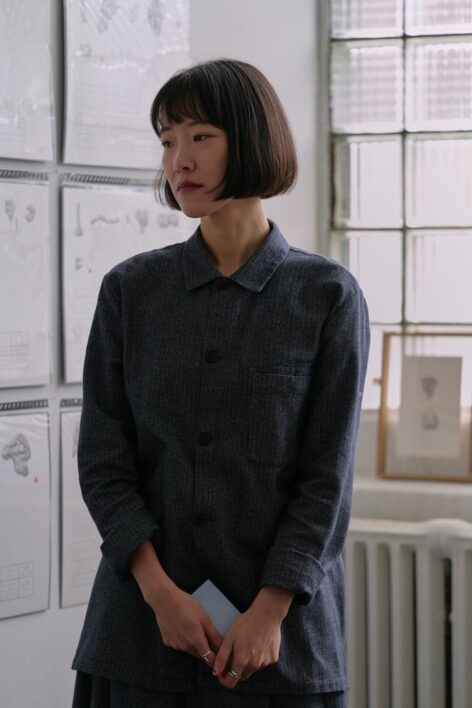 Yuchen Chang, in a gray button-up long-sleeve shirt, stands before a window and a white wall covered in white posters. Looking away from the camera, Yuchen drapes her arms in front of her low torso, with a small light blue rectangular object held between her hands. Photo by Junyan Hu.
