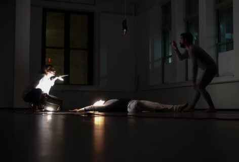 2019.11.19.Open Performance. Three dancers perform in a darkened studio. A dancer lays in the center. Another flashes a light in the first dancer's face. The third dancer controls an abject that hangs above the dancer laying down. Photo by Harry Shunyao Zhang.
