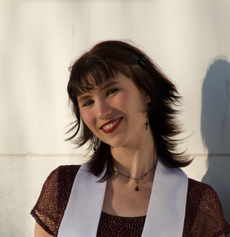 Alice is smiling directly at the camera, wearing a maroon dress with a white stole, with a white sunlit background. Photo by Guin MacLowry.