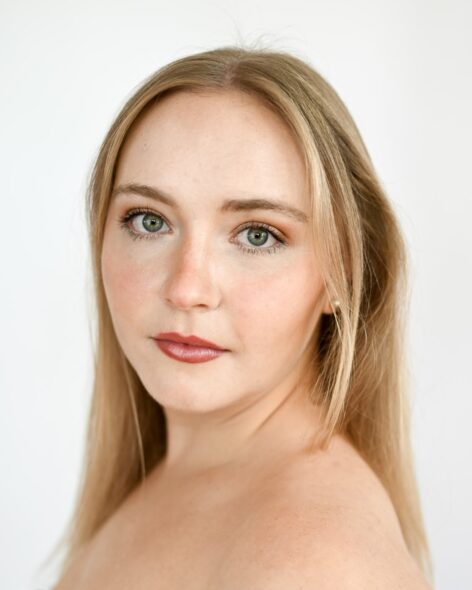 Photo of a young woman with long blonde hair looking over her shoulder in front of a plain background. Photo by Milan Furtado.