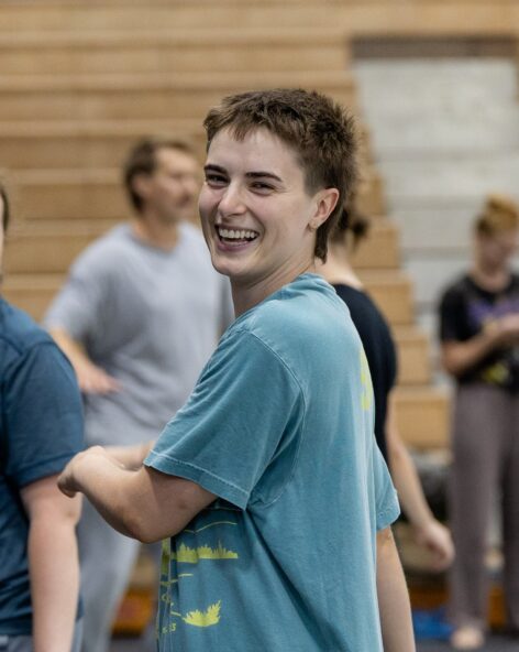 Dawn, a white non-binary person with short brown hair and a teal t-shirt on, is smiling towards the camera while walking to the left of the frame. Photo by Olivia Moon.