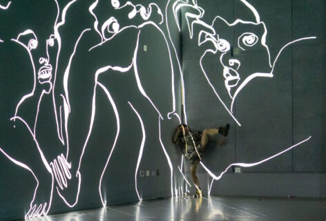 Jesse Zaritt, a white male dancer, moves within a projection of his animated line drawings. He leans against the corner of a stage space, one leg extended off the floor, surrounded by large drawn faces and arms.
