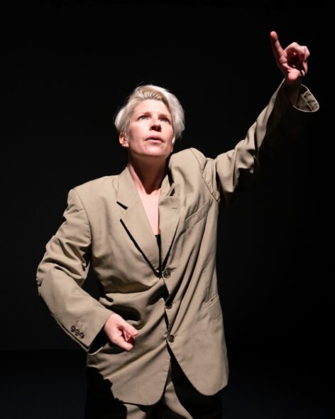 Close up image is of a bleached blond white woman with short hair wearing oversized tan suit pointing upwards with right hand. Left hand is in a held in loose fist by hip and gaze is upward and facial expression somewhat perplexed. Photo by Ellen Crane.