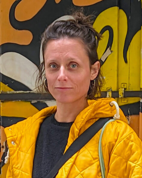 Portrait of Tina Valentan outdoors, wearing a bright yellow jacket and a black scarf, standing in front of a black-and-yellow graffiti-covered wall. She looks directly at the camera with a slight smile. Green foliage is visible in the background. Photo by Megajver.