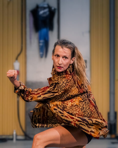 A female dancer is captured in a dynamic dance position. Her knees are bent, giving her body a low, grounded stance. She is wearing a long-sleeved leopard printed top, leaving her legs visible from the shorts down. Only one arm is clearly visible, bent at the elbow, while the rest of her body is turned partly away from the viewer. Her posture suggests movement and concentration, as if she has been caught in the middle of a dance. photo by Manoj Bandaru.